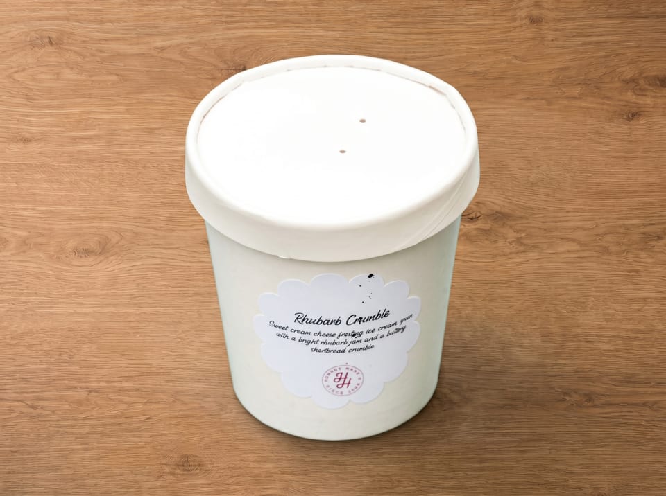 A white pint of "Rhubarb Crumble" ice cream with a decorative label on a wooden surface.