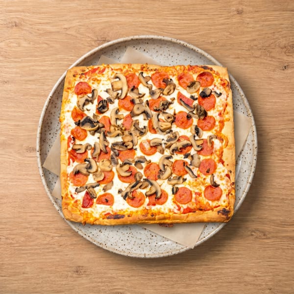 Square pepperoni and mushroom pizza on a speckled white plate, on a light wooden table.